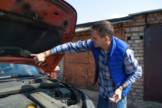 Man pointing at the engine under an open hood of an orange car in a garage driveway, checking problem, auto repair, vehicle maintenance, diy service.