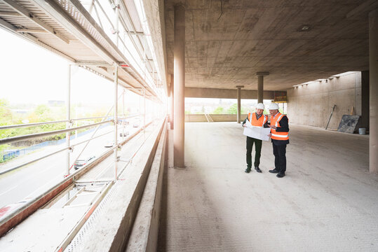 Two men with plan wearing safety vests talking in building under construction