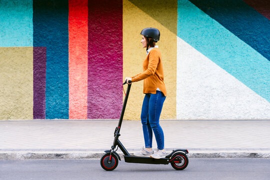 Woman wearing helmet and riding electric push scooter in front of multi colored wall