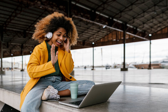 Cheerful Afro woman with in-ear headphones waving hand during video call through laptop in seating area