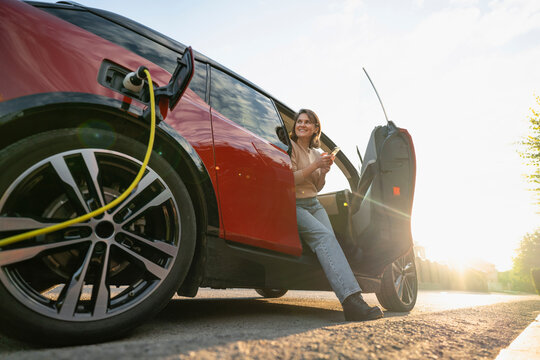 Smiling woman sitting with mobile phone and charging car at roadside