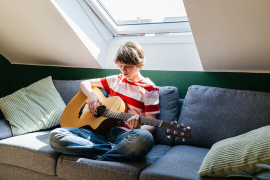 Girl playing acoustic guitar on a sofa at home on a sunny day