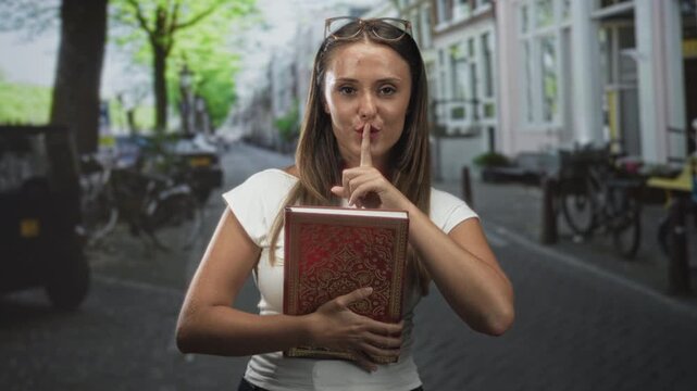 Woman holding a leatherbound red book against her chest, finger to lips for silence on a street with bicycles and buildings; secrecy privacy study.