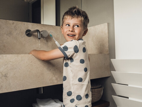 A smiling child brushes teeth in a natural stone bathroom