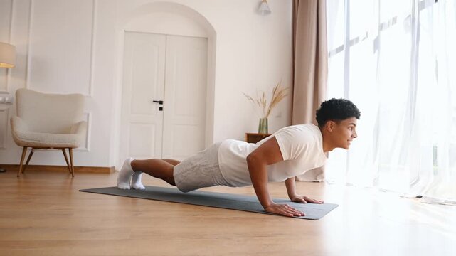 Young man performs a knee push-up on a yoga mat in a sunlit living room, focusing on strength, form and core engagement during a convenient at-home fitness routine for beginners and active lifestyles.