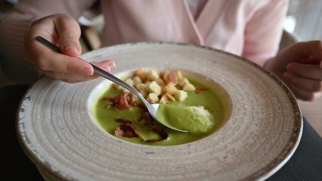 Woman eating delicious creamy green vegetable soup with bacon and crunchy croutons in restaurant.
