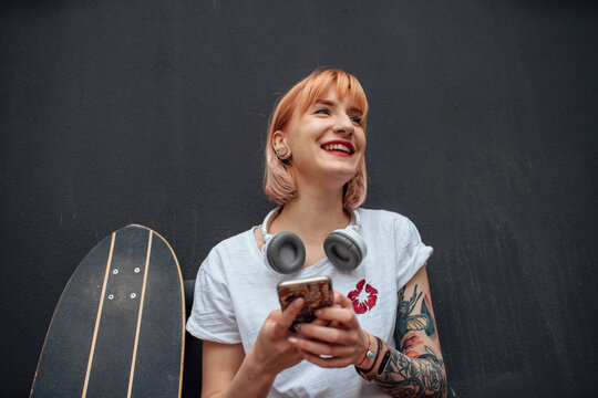 Happy young woman with skateboard, headphones and cell phone