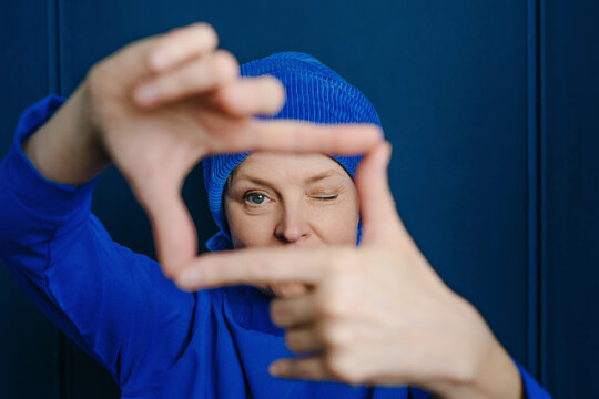 Playful woman in beanie making finger frame on blue wall
