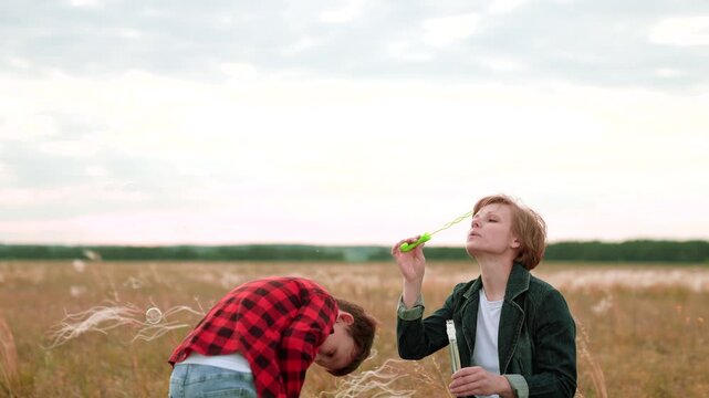 Woman joyfully blows bubbles while son catches bubbles on field. Mother with boy playing with bubbles in nature at sunset. Woman with son shares laughter and fun while chasing bubbles