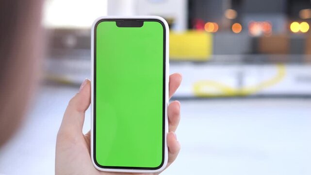 Hand holds smartphone with green screen. Background blurred with warm lights. Fingers tap screen gently. Ideal for app demos, digital marketing, or UI design. Suitable for commercial use in tech