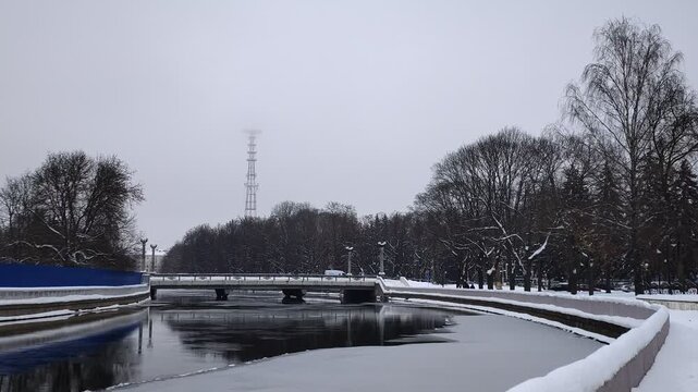 A partially ice-covered canal with concrete banks and a parapet runs between the trees. Snow covers the ground. There is a bridge over the water nearby. Cars are driving. Cloudy winter weather