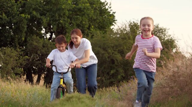 Smiling girl runs while mother teaches little boy to ride bicycle on grassy rural road at sunset. Mother with girl and boy practice bicycle riding in field. Caring mother with boy and girl on vacation