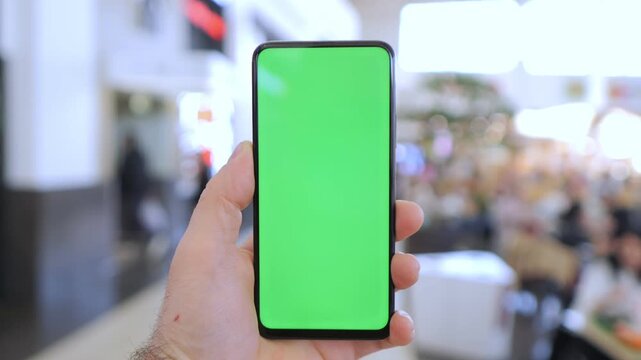 Hand holds smartphone with green screen. Background shows busy airport lounge. People move around casually. Good for app demos or travel-related content. Ideal for mockups or digital interface design