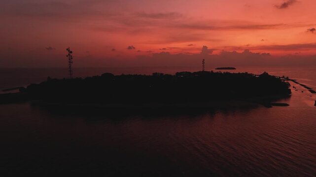 Aerial view of the silhouette of Kendhoo island with communication towers under a vibrant red sunset sky over the calm Indian Ocean in Kendhoo, Baa Atoll, Maldives.