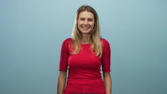 Woman blonde and young smiling and laughing with hand covering face, wearing red top and jeans while standing against a pale blue studio backdrop; joy playfulness.