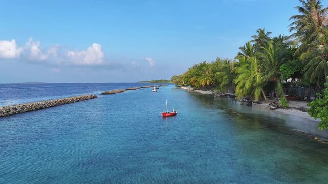 Aerial view of a tropical coastline with turquoise water, a stone breakwater, and palm trees along the shore in Kendhoo, Baa Atoll, Maldives.