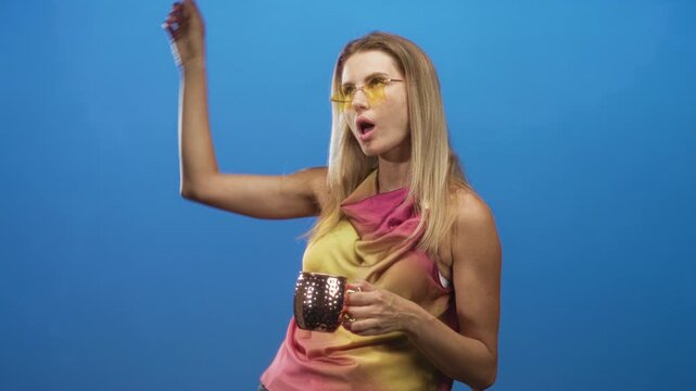 Blonde woman holding a copper cocktail mug, bare right arm raised to forehead and puckering lips against a blue studio backdrop; playful.