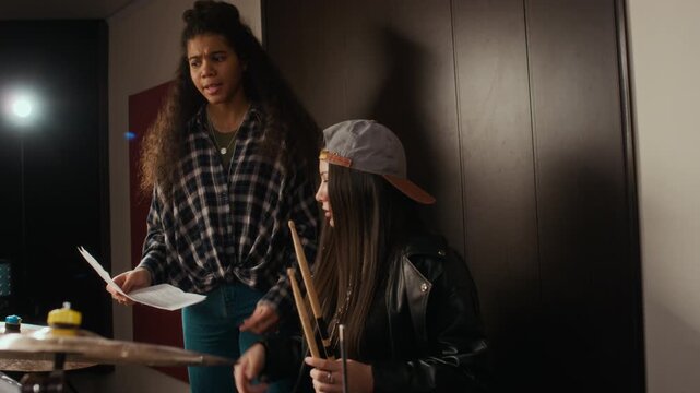 Caucasian female teenager practicing drums while African American female teenager holding sheet music explaining rhythm during rehearsal. Clip is perfect for music school advertising