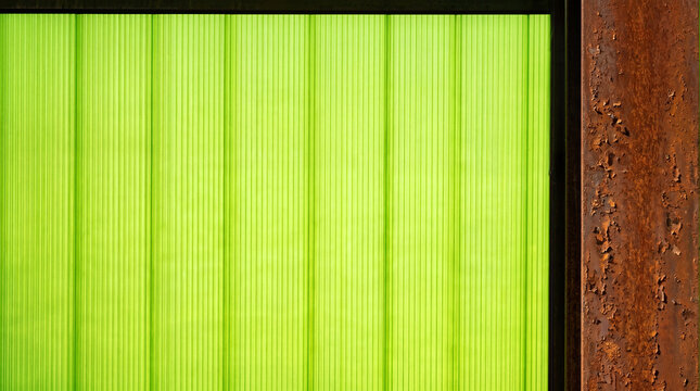 Translucent green corrugated plastic panel framed by weathered rusty metal. Abstract architectural detail showing contrast between modern synthetic material and decayed industrial texture.