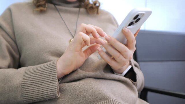 Person in cozy sweater uses smartphone while seated. Fingers scroll or tap on screen. Calm and focused, perhaps during travel or downtime. Great for tech, communication, or modern lifestyle concepts