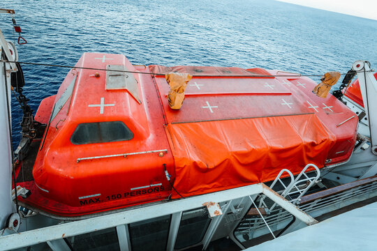 High angle view of an orange enclosed emergency lifeboat marked for maximum 150 persons, mounted on a passenger vessel deck with the blue sea in the background.