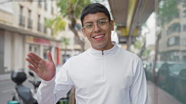 Man waving right hand and smiling, wearing glasses and white zip pullover while standing on an urban sidewalk street with parked scooters; friendly greeting.