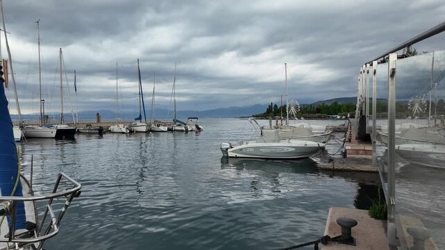 Bardolino, Italy - May 14, 2026 - Sailboats docked in a serene harbor under a cloudy sky with distant mountains