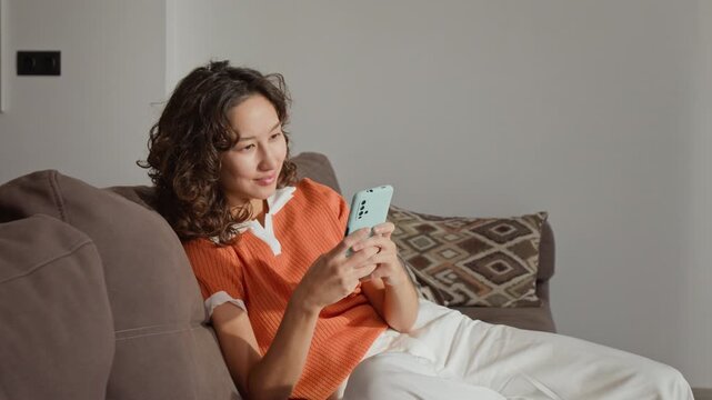 Asian young adult woman sitting on sofa and using smartphone at home. Clip is suitable for mobile application marketing, social media campaigns, and digital lifestyle advertisements