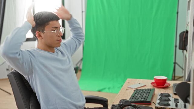 Young man photographer leaning back with hands behind head beside camera and keyboard in studio with green screen; calm reflection.