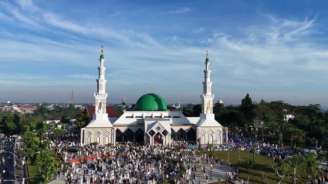 An aerial view of the Baitul Faizin Grand Mosque during the busy Eid al-Fitr prayer, with the signature green dome and twin minarets, a vast congregation on blue mats