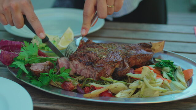 Hands slicing grilled bone in steak at outdoor restaurant table capturing shared dining experience, gourmet cuisine and authentic social mealtime atmosphere