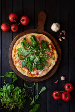 homemade prosciutto, arugula and parmesan pizza on a wooden table sorunded by ingridients on a darck background