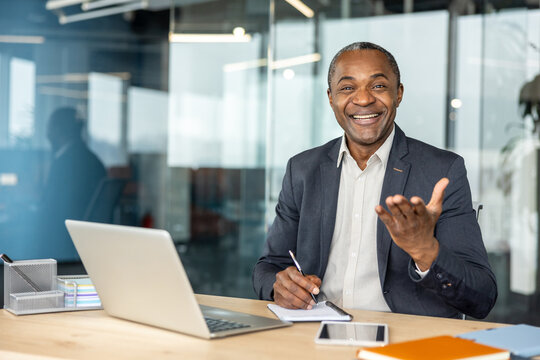 Mature businessman laughing and gesticulating while sitting at a desk with a laptop and notebook in a modern office setup, representing successful communication and leadership