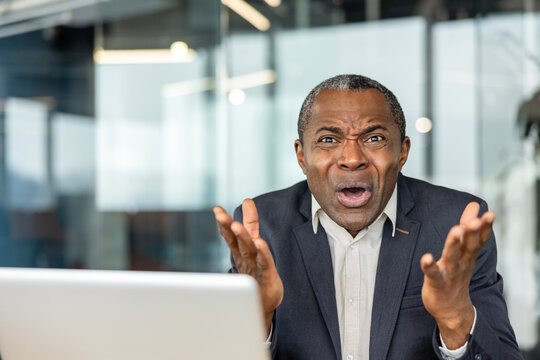Mature adult businessman expressing intense shock, surprise, and dissatisfaction while raising hands in frustration, reacting to bad news or a problem on a laptop screen in an office setting