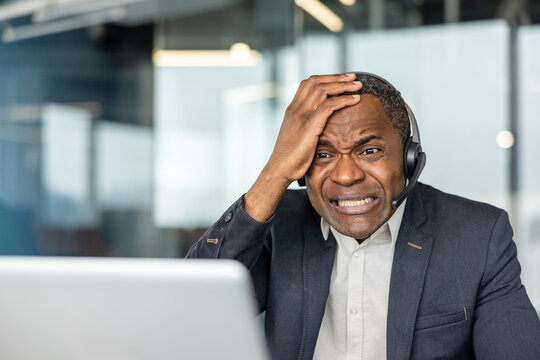 Frustrated mature employee wearing a headset and suit holding his head, experiencing stress and a difficult situation while working on a laptop at his office desk