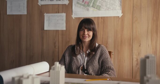 A designer sits at a table with plans and a building model while discussing work in a studio with natural light