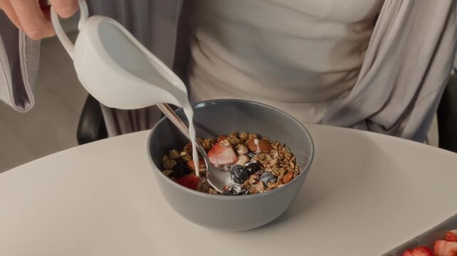 Close up on hands of young adult woman pouring milk from pitcher into bowl of granola with fresh berries and stirring breakfast. Clip perfect for healthy lifestyle and organic food commercials