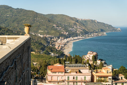 View of the Mediterranean coastline with colorful buildings, lush green hills, and a sandy beach from a high vantage point Taormina, Sicily, Italy.