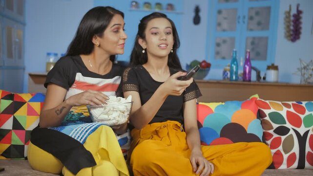 Mother-daughter enjoying their favorite program on television - leisure moment. Medium shot of two family members relaxing at home watching TV while eating wafers / chips - happy urban lifestyle