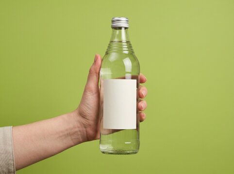 Transparent Glass Bottle with Ribbed Neck and Blank Label Held in Hand Against Green Background