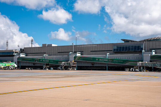 Ground view of O. R. Tambo International Airport in Johannesburg, green jet bridges extend from a modern concourse, service vehicles sit parked under a bright sky.