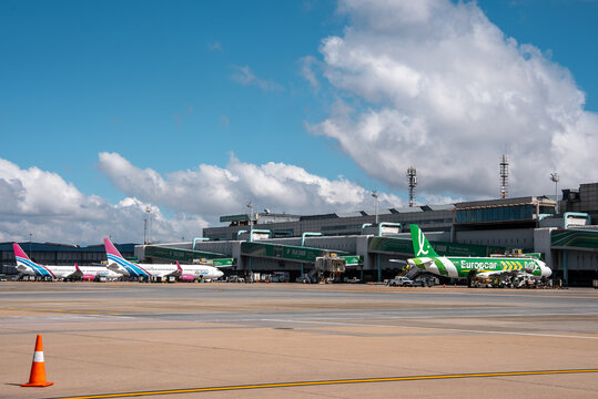 O. R. Tambo International Airport apron shows multiple jets at green jet bridges, a vivid green Europcar aircraft, low modern terminals with radio masts, and wide taxiways.