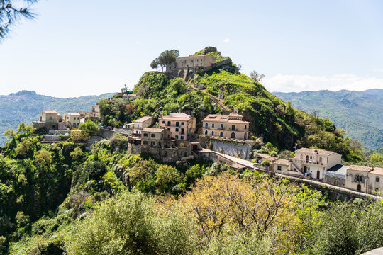 View of the historic hilltop village of Savoca with traditional stone houses nestled on a lush green mountain ridge under a clear blue sky Savoca, Sicily, Italy.