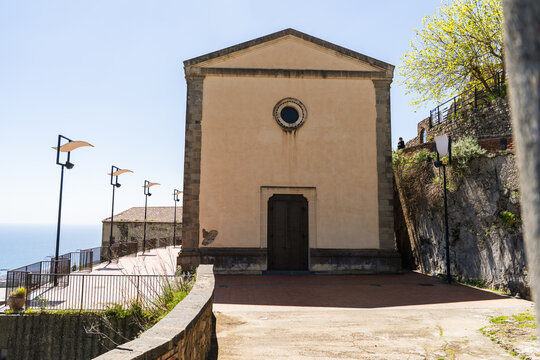 View of the historic Chiesa di San Michele with its simple facade and circular window on a stone terrace overlooking the sea in Savoca, Sicily, Italy.