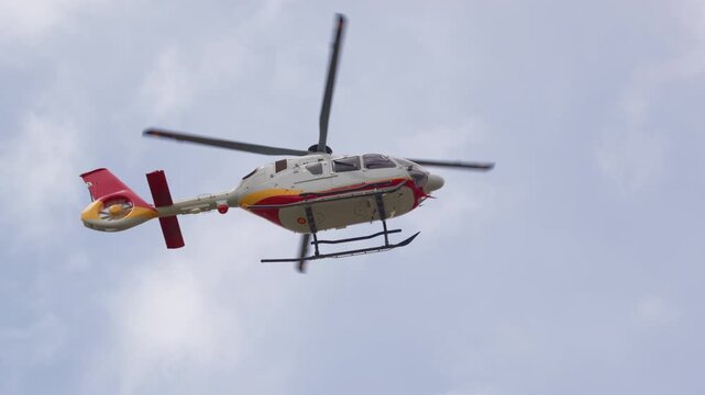 A red white and yellow helicopter with twin landing skids flies in profile across a soft cloudy sky, with the main rotor blades spinning above the cabin