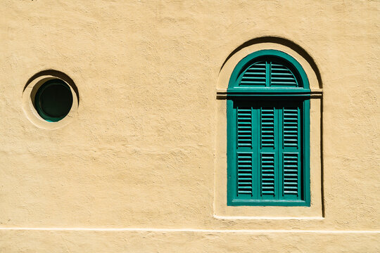 View of traditional green wooden shutters on an arched window and a small circular window set in a textured yellow stone wall in Rabat, Western Region, Malta.
