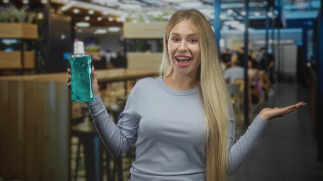 Woman presenting mouthwash bottle with open mouth and palm up gesture in a cafe dining area, smiling at camera; cheerful promotion.