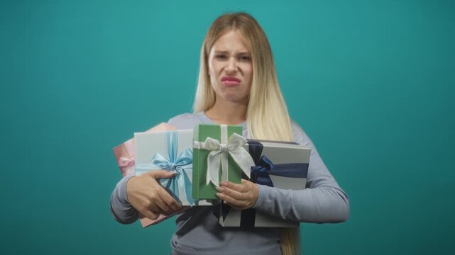 Woman clutching multiple wrapped gift boxes, grimacing with visible face and hands against teal studio backdrop; disgust.