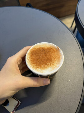 View of a hand holding a paper cup of coffee with cinnamon powder on top at a cafe table in Beylikduzu, Istanbul, Turkey.