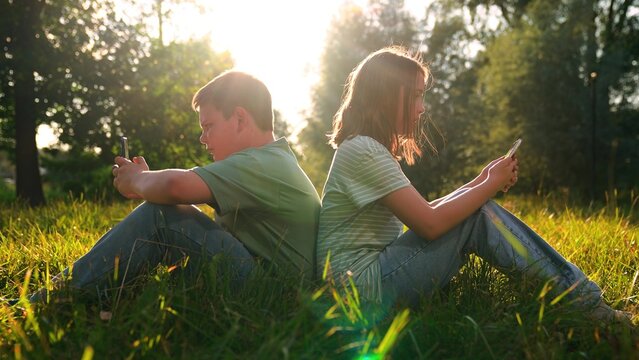 Children relaxing in park with phones, Side-by-side but online apart, Nature backdrop for digital life, Golden light smartphone moment, Social feed in the fresh air, Rural meadow tech contrast